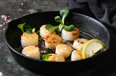 Fried scallops with butter lemon spicy sauce in cast-iron pan served with green salad and textile napkin over old dark metal background.
